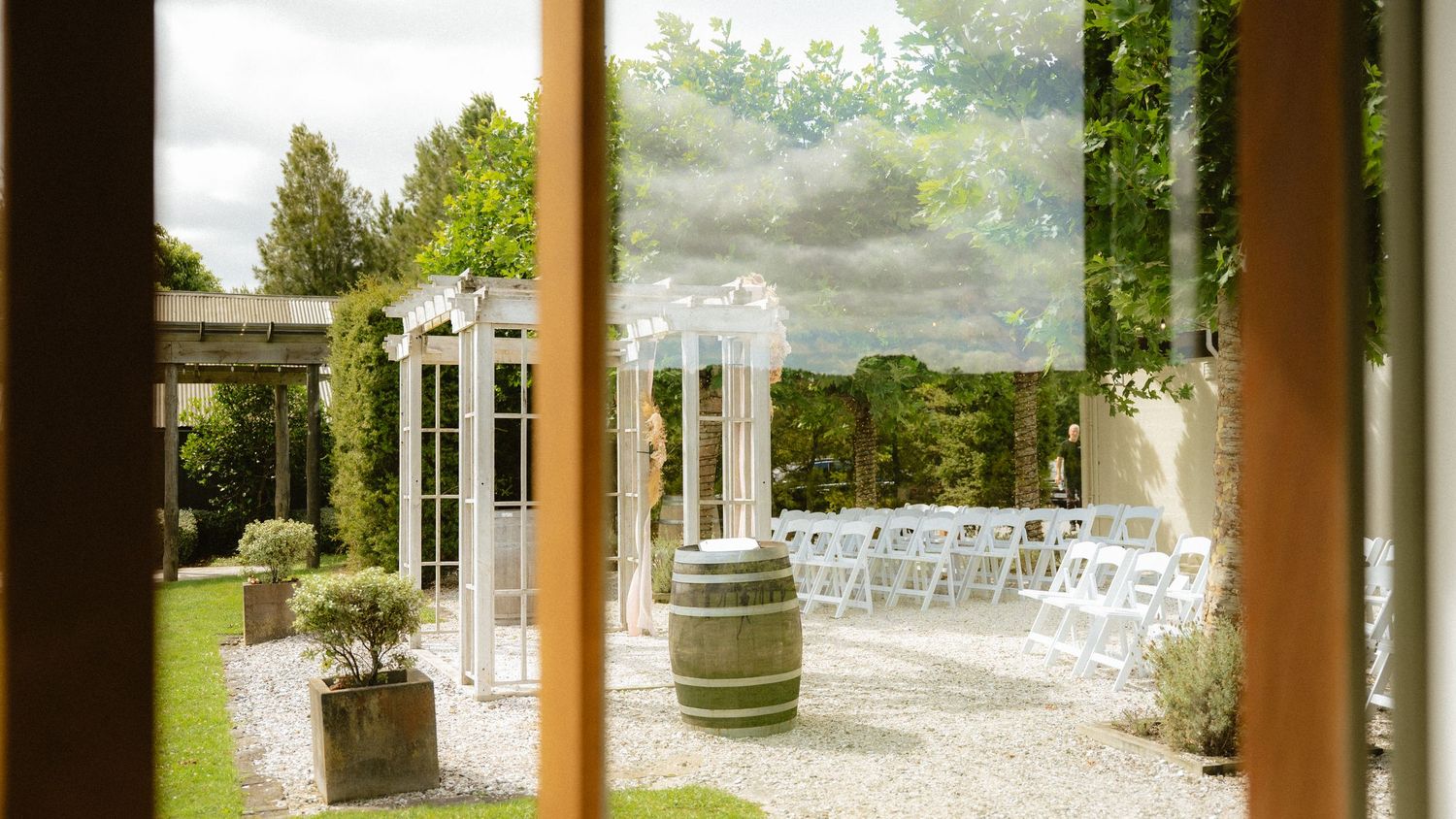 Elegant outdoor wedding ceremony setup with white chairs, barrel, and arbor viewed through open wooden doors.