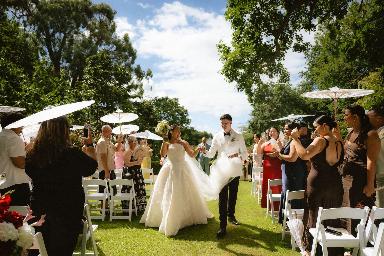 Bride and groom walk down outdoor wedding aisle lined with guests holding white parasols on a sunny day.