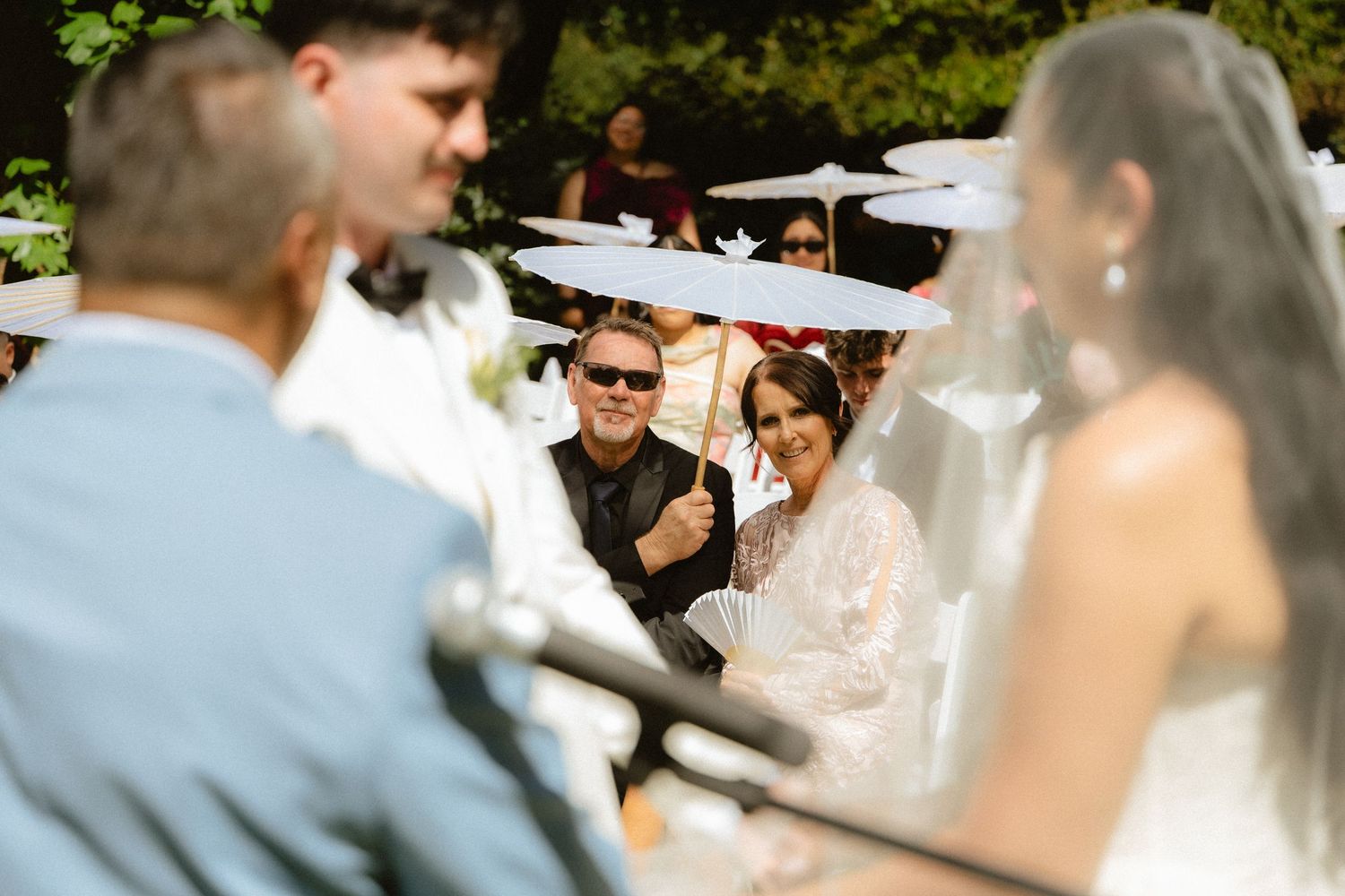 Outdoor wedding ceremony with smiling bride and groom exchanging vows among guests holding white parasols.