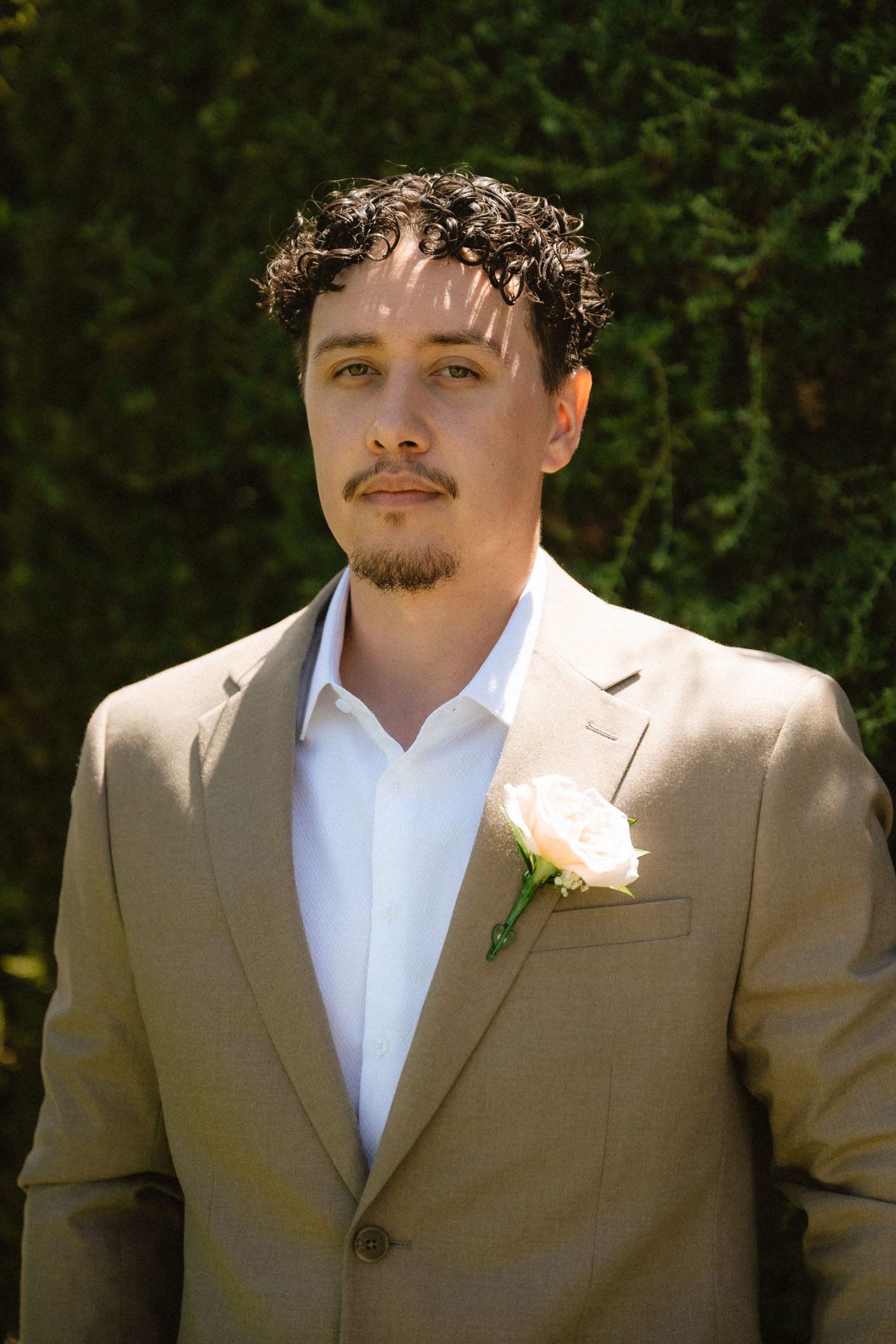 Young man in tan suit with white rose boutonniere standing outdoors against green foliage backdrop.