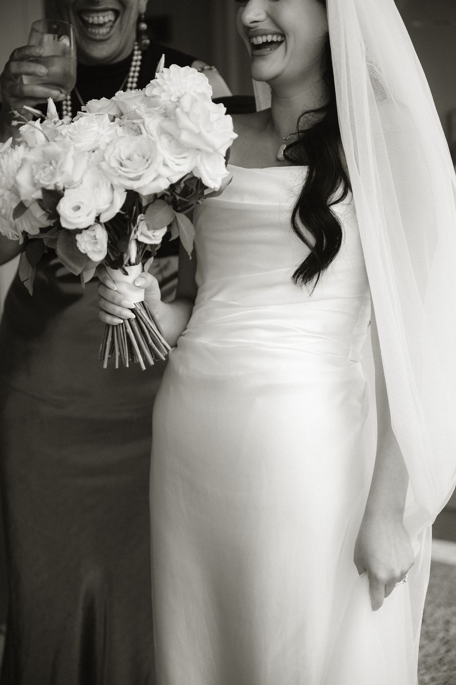 Black and white photo of a smiling bride in a white gown holding a rose bouquet with a woman beside her.