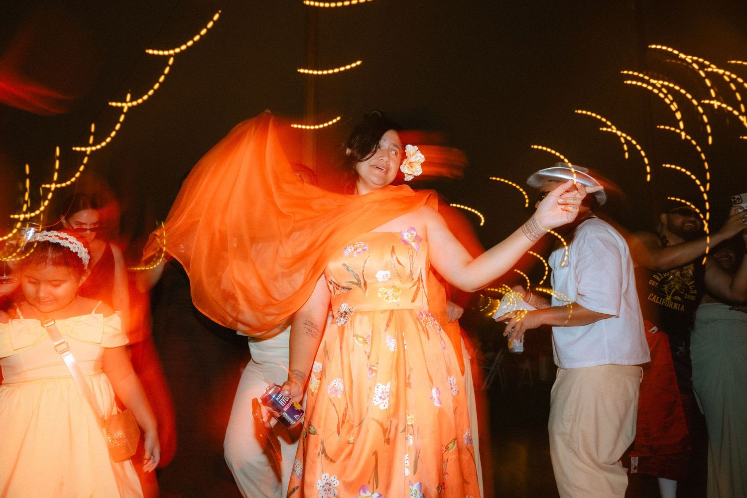 Woman in flowing orange dress dancing joyfully at a festive evening celebration with colorful lights swirling in background.