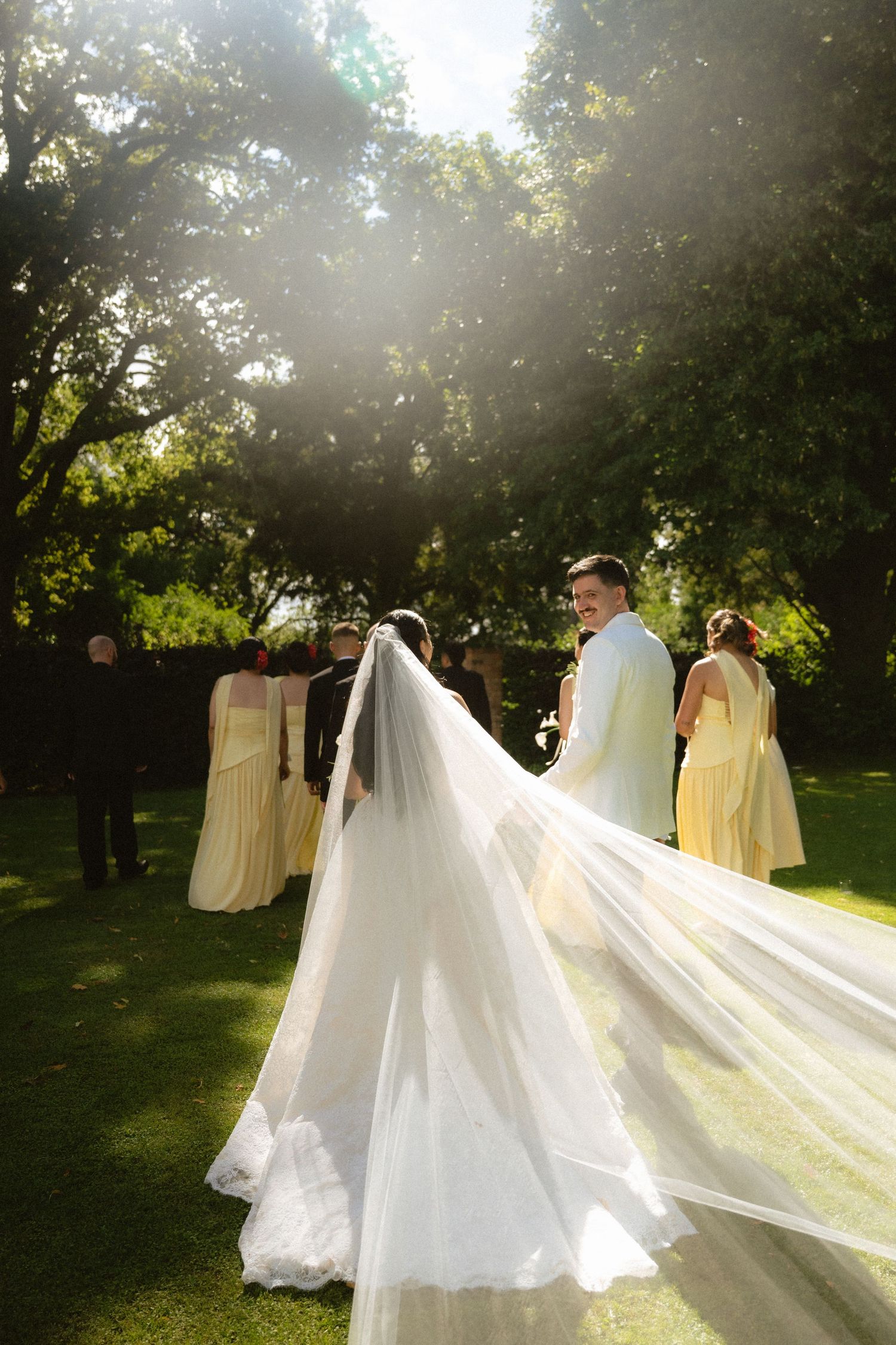 Bride and groom share a romantic outdoor wedding moment, with flowing veil catching sunlight, bridesmaids in yellow dresses nearby.