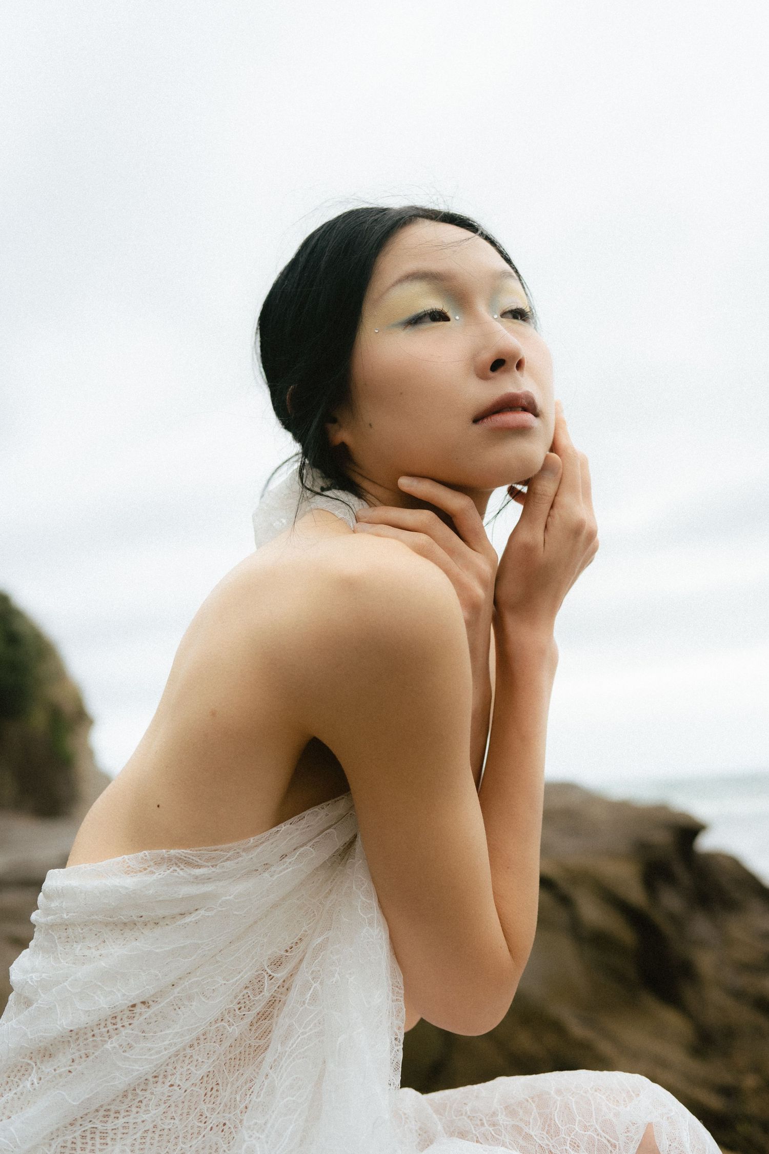 Young Asian woman wrapped in white lace fabric poses thoughtfully on rocky beach with overcast sky.