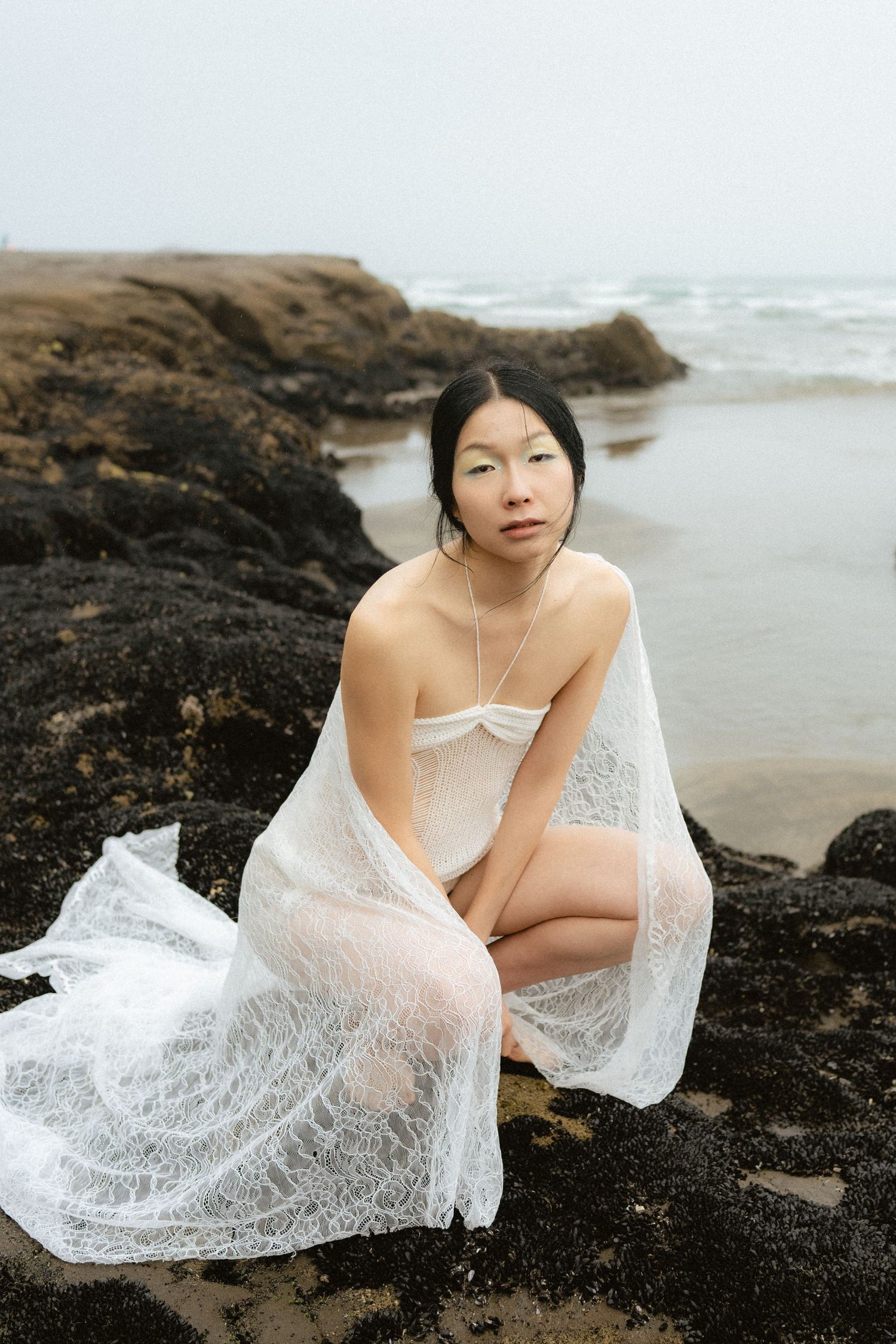Woman in white lace fabric sitting on rocky shoreline with ocean waves and misty sky in background.
