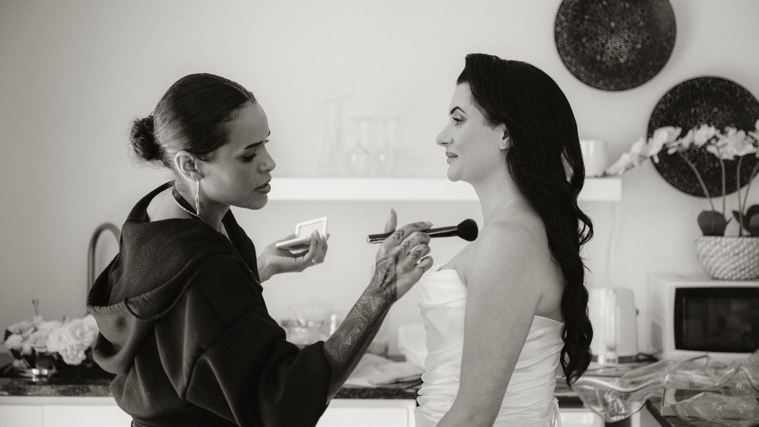 Black and white photo of a makeup artist applying blush to a bride in a strapless wedding dress before her ceremony.