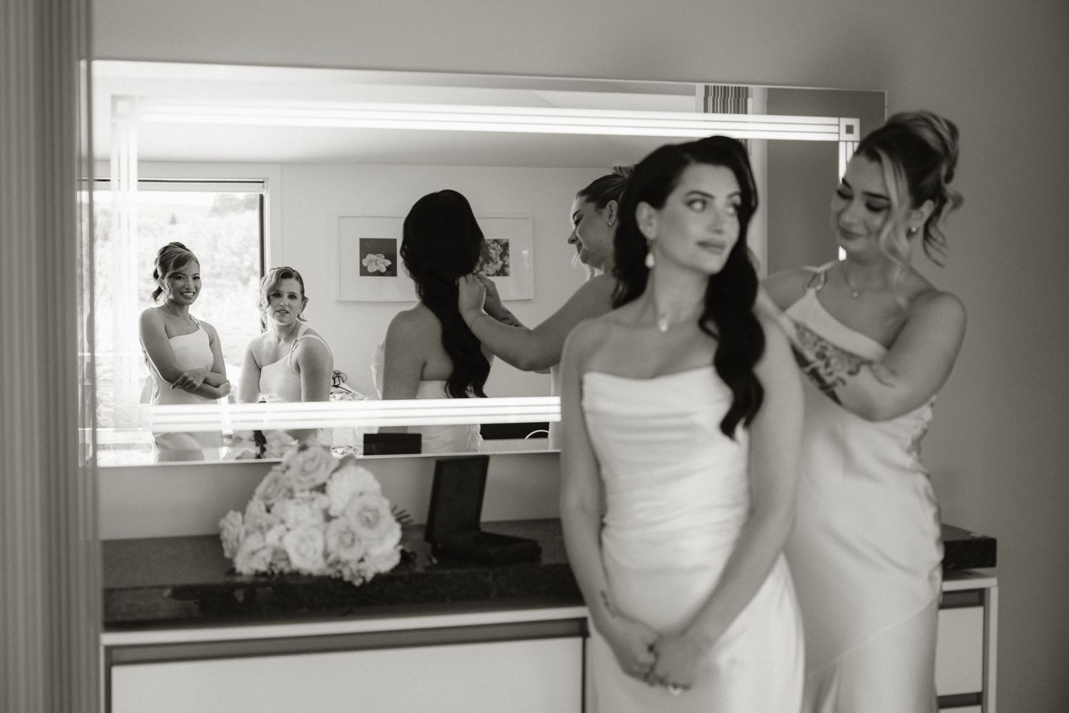 Black and white photo of bride getting ready with bridesmaids adjusting her dress and hair near a lit vanity mirror.