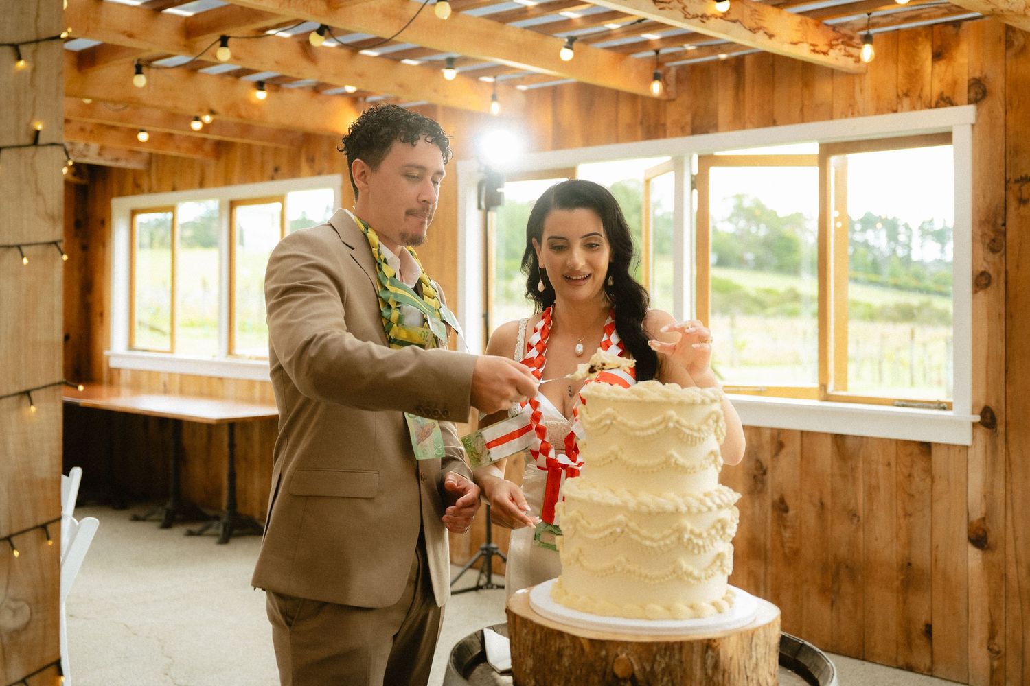 Bride and groom cutting a rustic white wedding cake on a wood stump in a warm wooden venue.