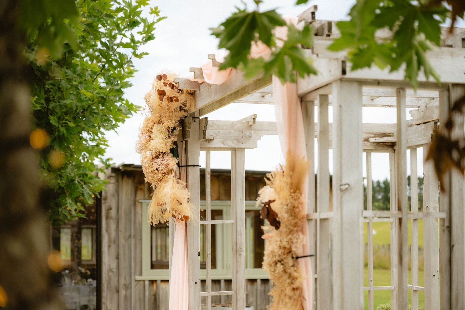 Rustic white wooden wedding arch adorned with blush pink ribbons and dried floral arrangements outdoors.