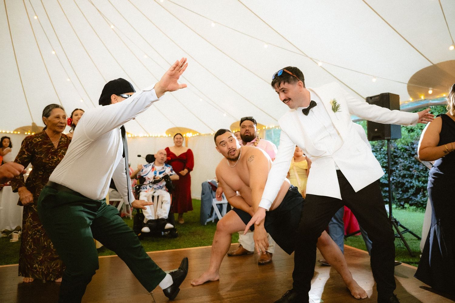 Men dancing energetically at a wedding reception inside a marquee tent, with guests watching in the background.