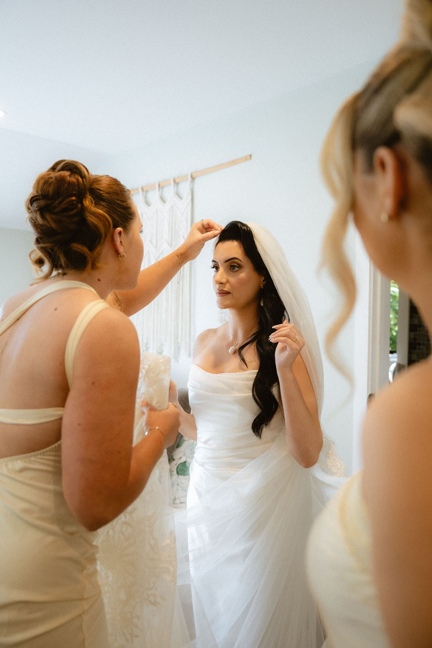 Bride in white strapless gown gets veil adjusted by bridesmaid in cream dress before wedding ceremony.