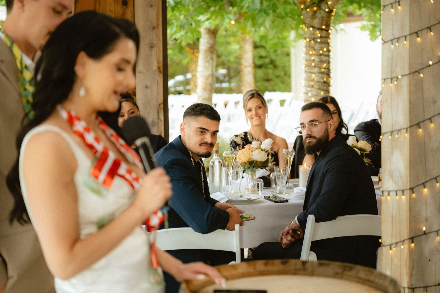 Wedding reception guests smiling and laughing at decorated outdoor venue with string lights and floral centerpieces.