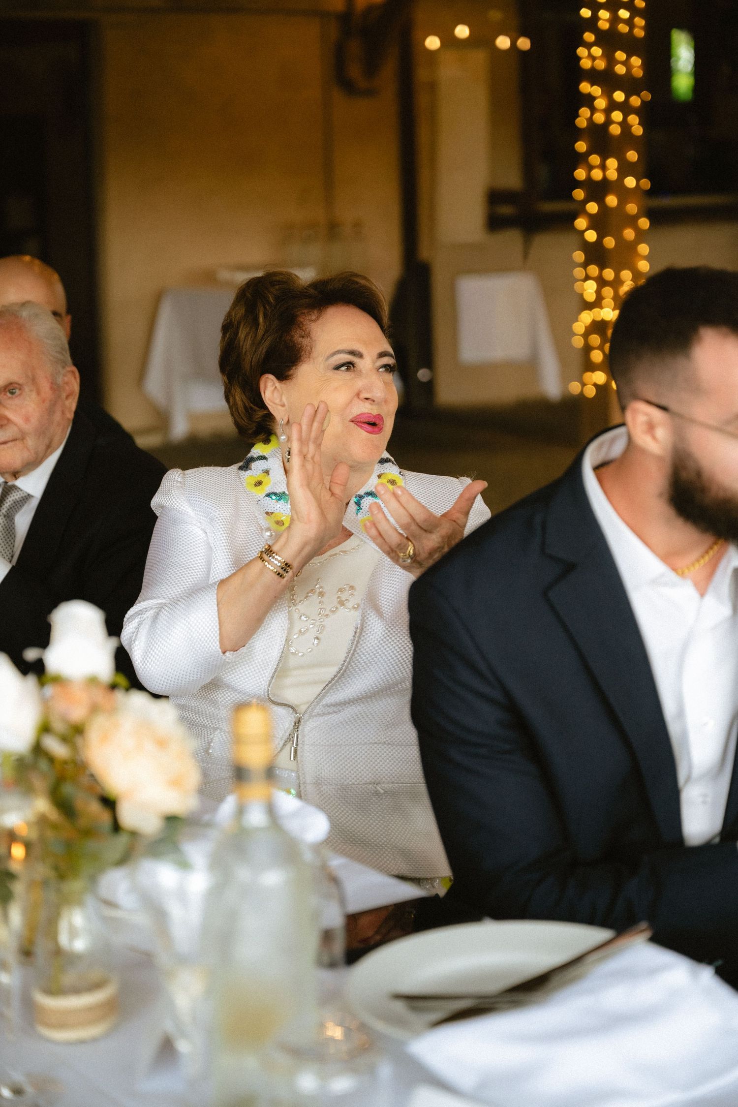 Elegant woman in white outfit clapping at a formal wedding reception dinner with bokeh lights in background.