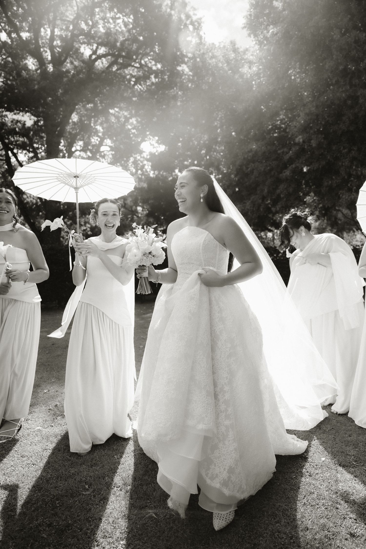 Black and white photo of a radiant bride in a lace gown walking with bridesmaids holding parasol outdoors.