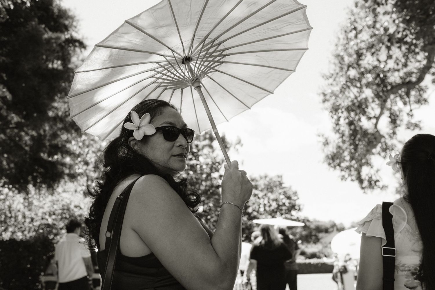 A woman wearing sunglasses and a flower in her hair holds a parasol umbrella at an outdoor garden event.