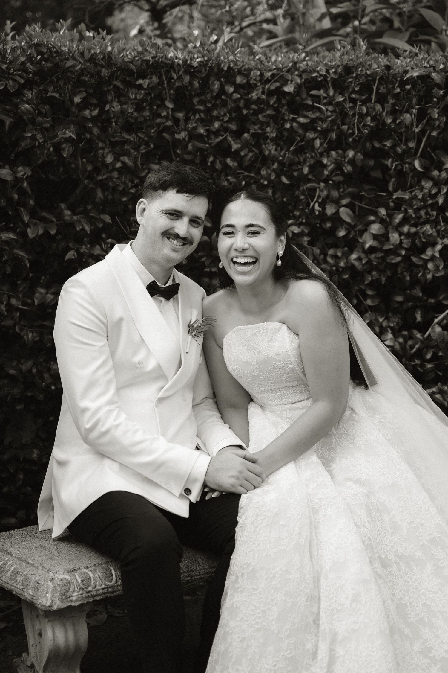 Happy bride and groom in black and white wedding portrait, sitting on bench surrounded by lush greenery.