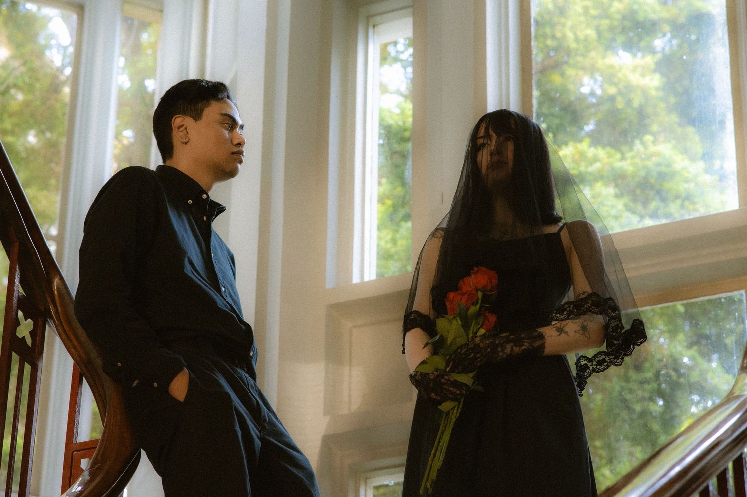 A man in black suit and woman in black dress with veil holding red roses stand on elegant staircase.