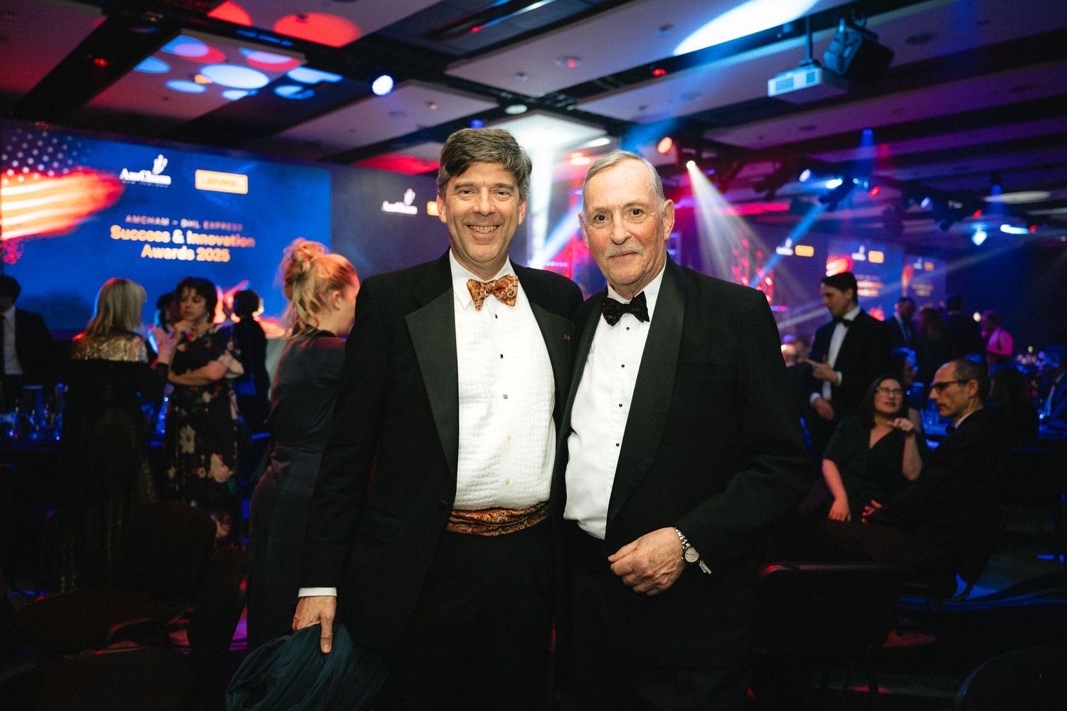 People in formal attire gather at an evening event with blue lighting and American flags visible in the background.