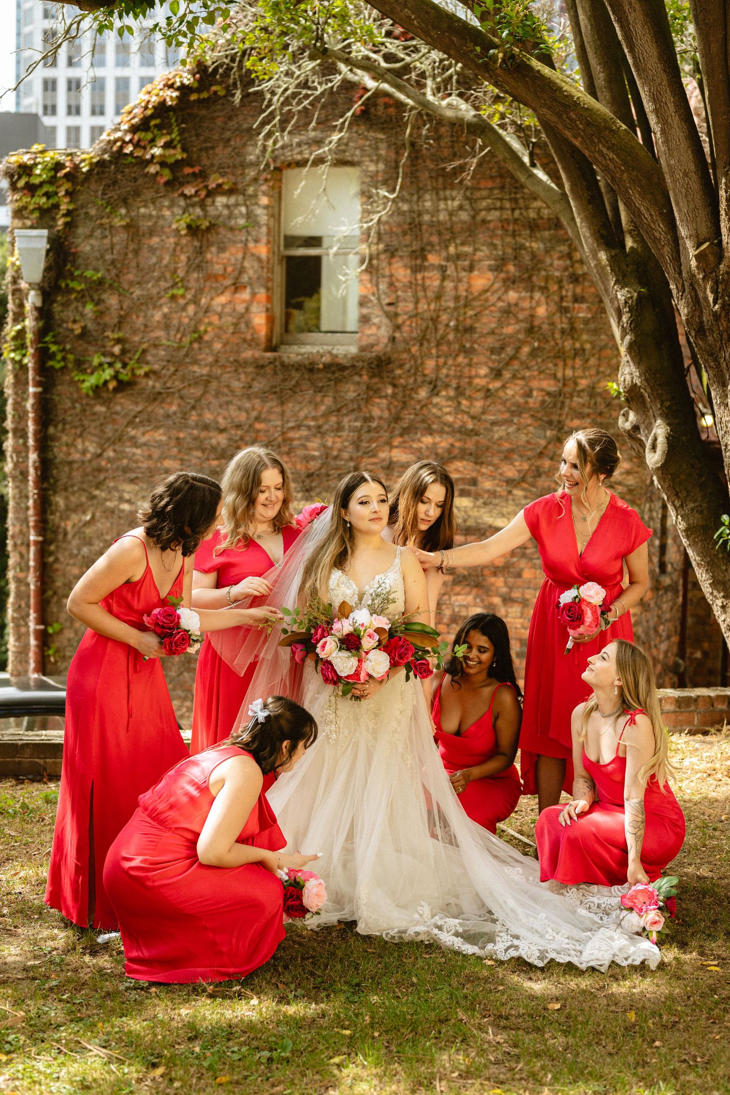 Bridal party in red dresses poses creatively on grass near a brick wall during outdoor photo session.