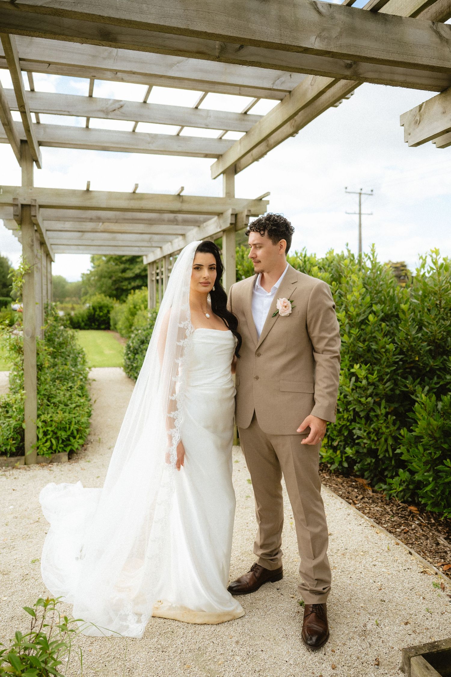 Bride in white gown with veil and groom in tan suit pose together on a garden path under a wooden pergola.