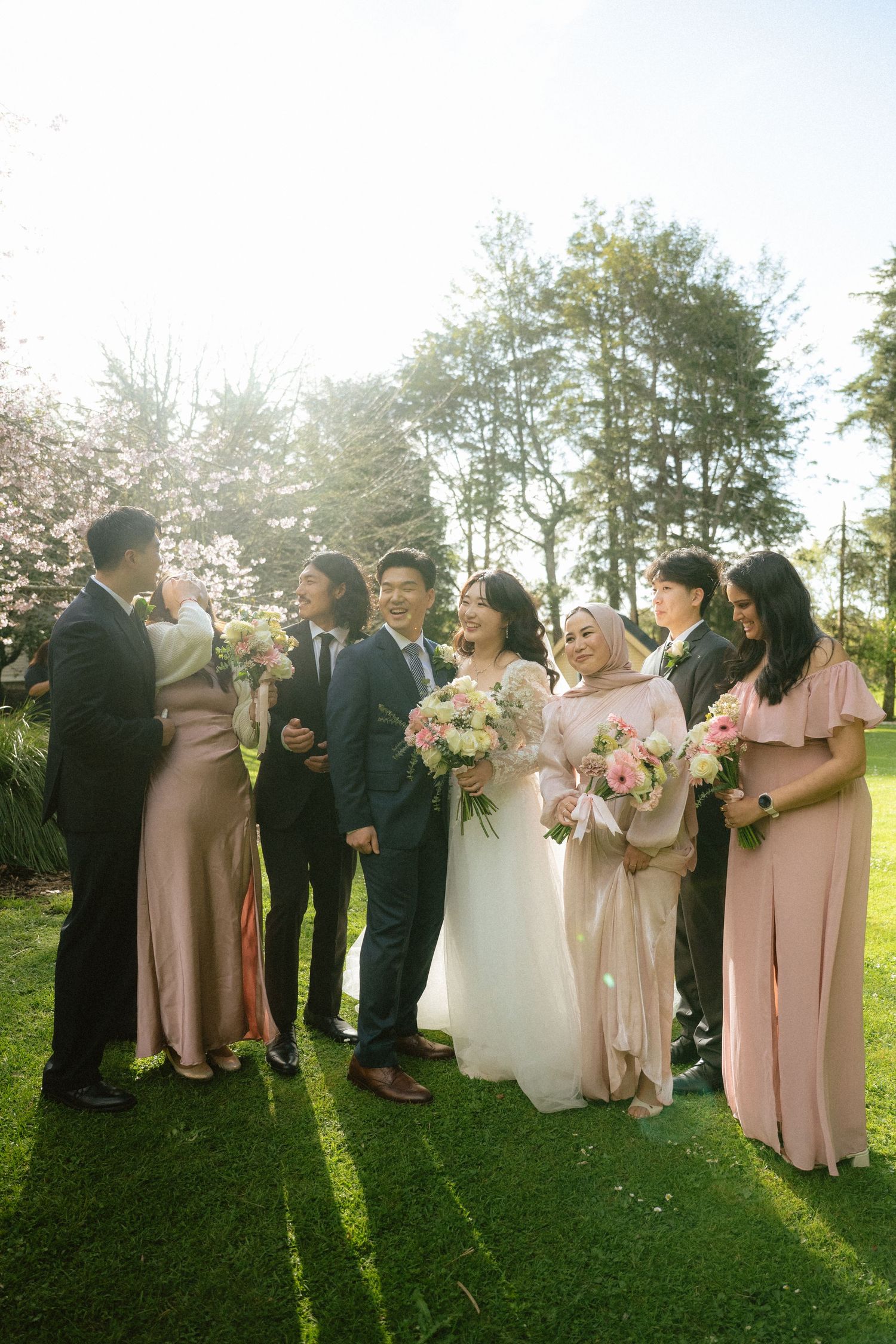 Wedding party stands together on green lawn during golden hour with bridesmaids in pink dresses and sunlit trees behind them.