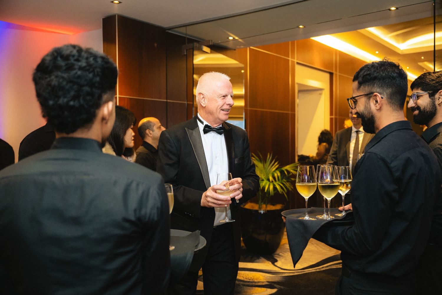 People in formal attire socializing at an elegant evening event with warm lighting and modern interior decor.