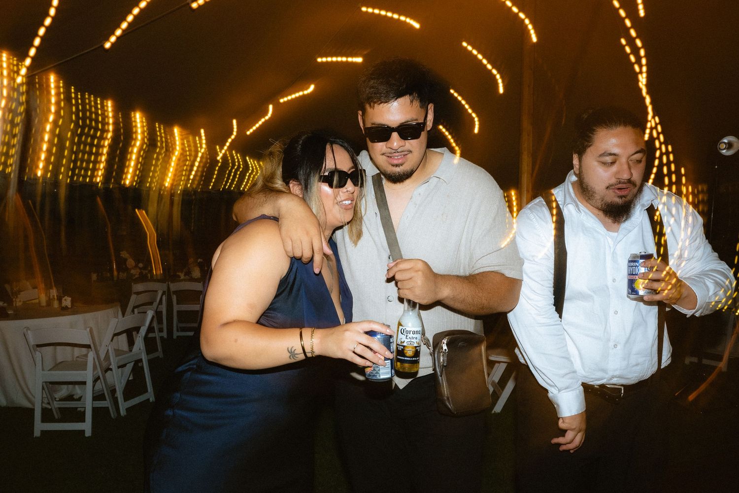 Three guests in formal attire pose and smile under glowing string lights at an elegant outdoor wedding reception.