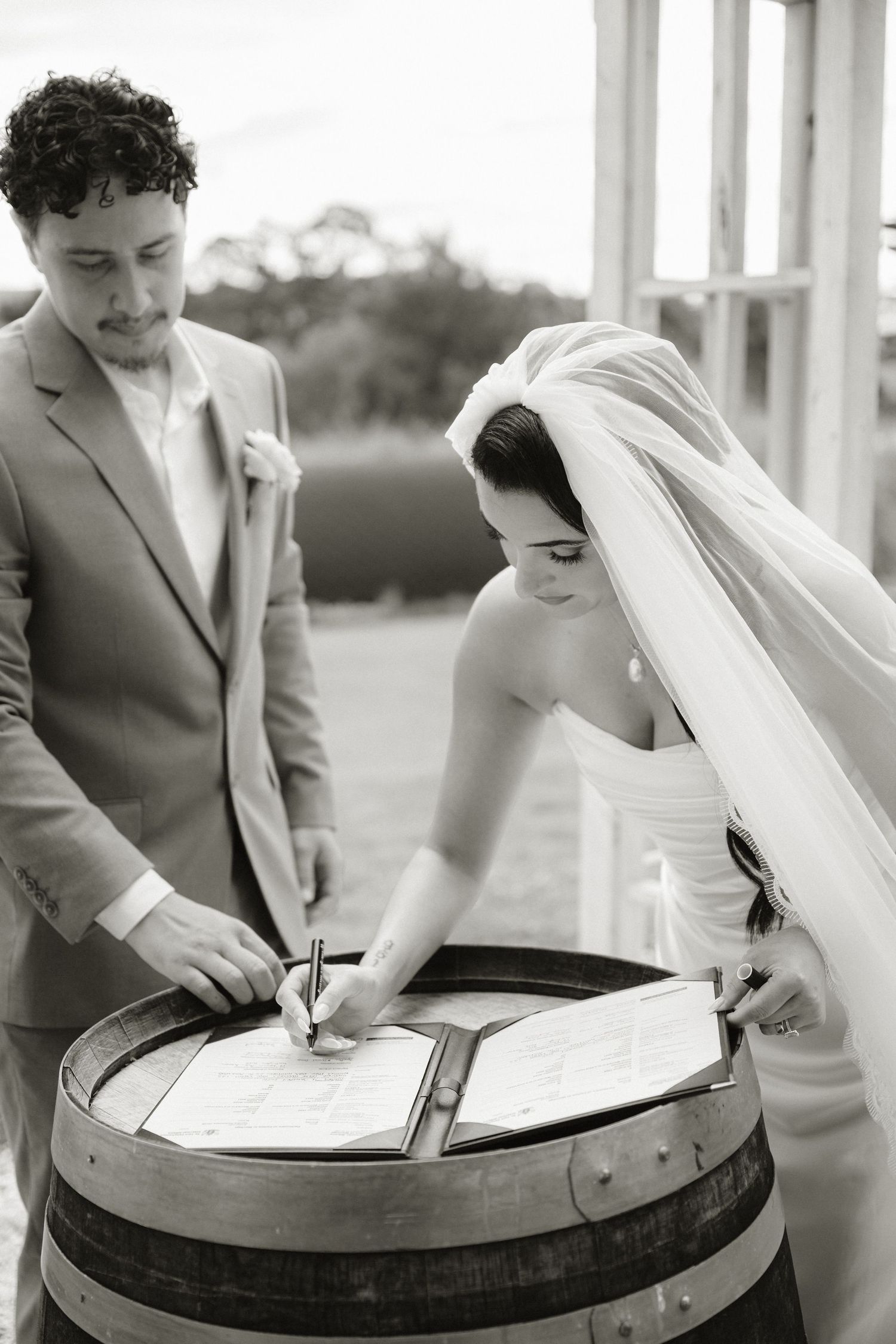 Bride signing wedding registry on a wooden barrel while groom watches during outdoor ceremony, black and white photo.
