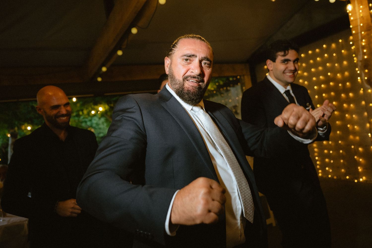Man in suit dancing energetically at an elegant evening wedding reception with string lights in background.