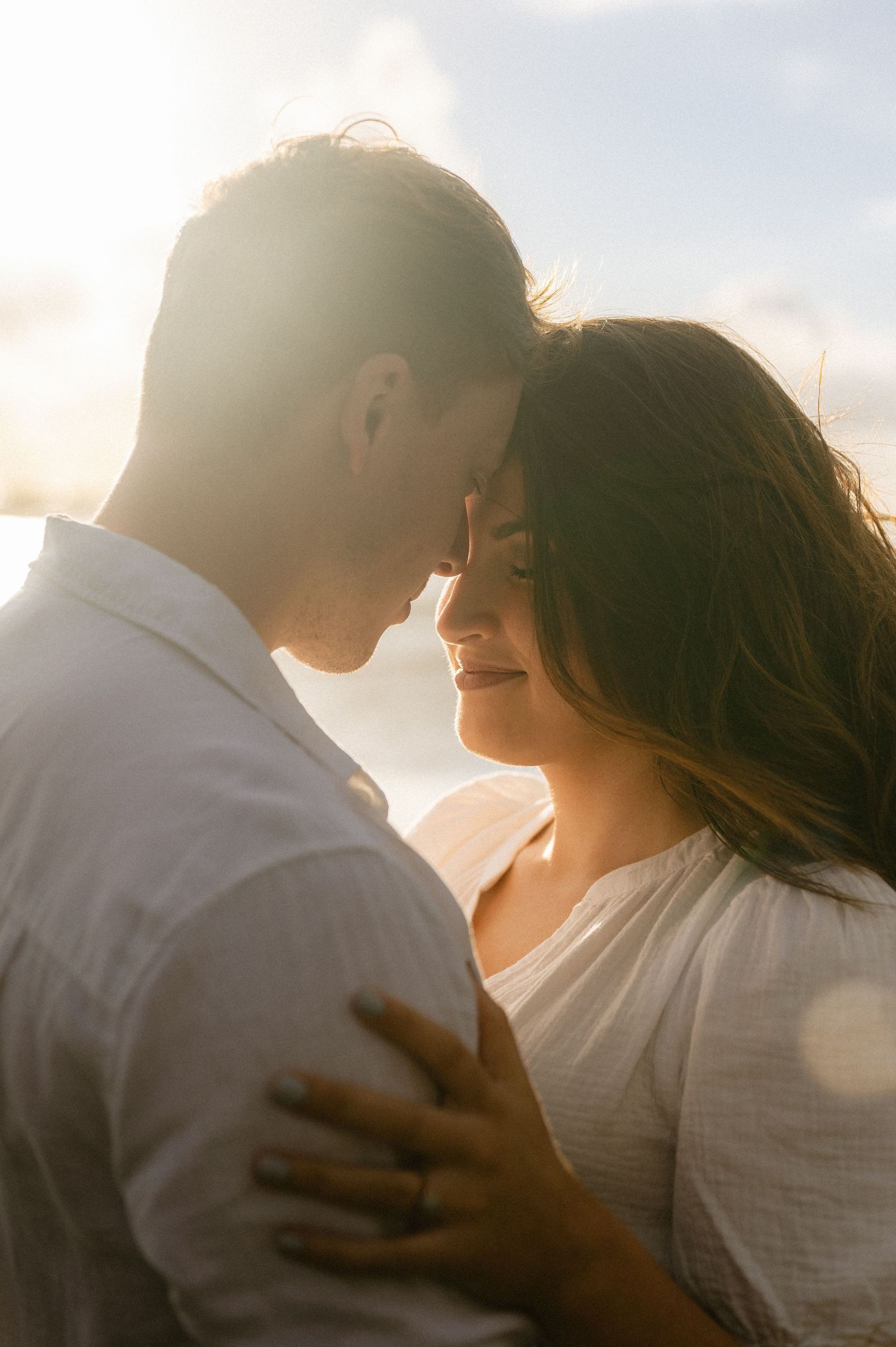 Romantic couple embraces in warm golden sunlight during sunset creating silhouettes against bright sky.
