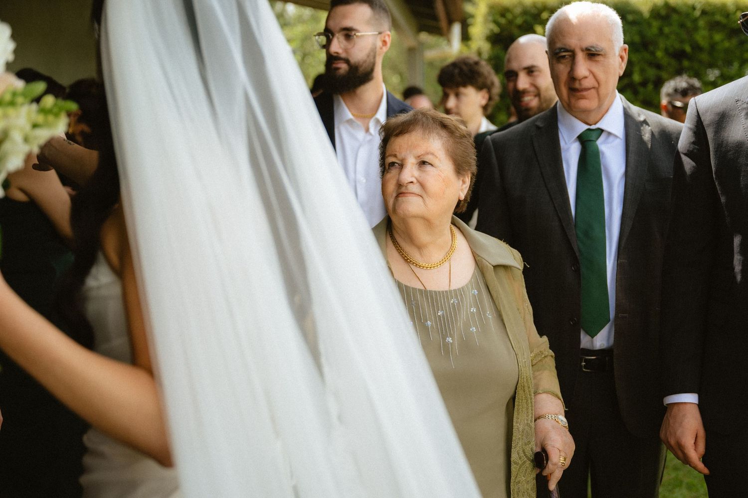 Elderly couple watching bride in white veil and gown at outdoor wedding ceremony.