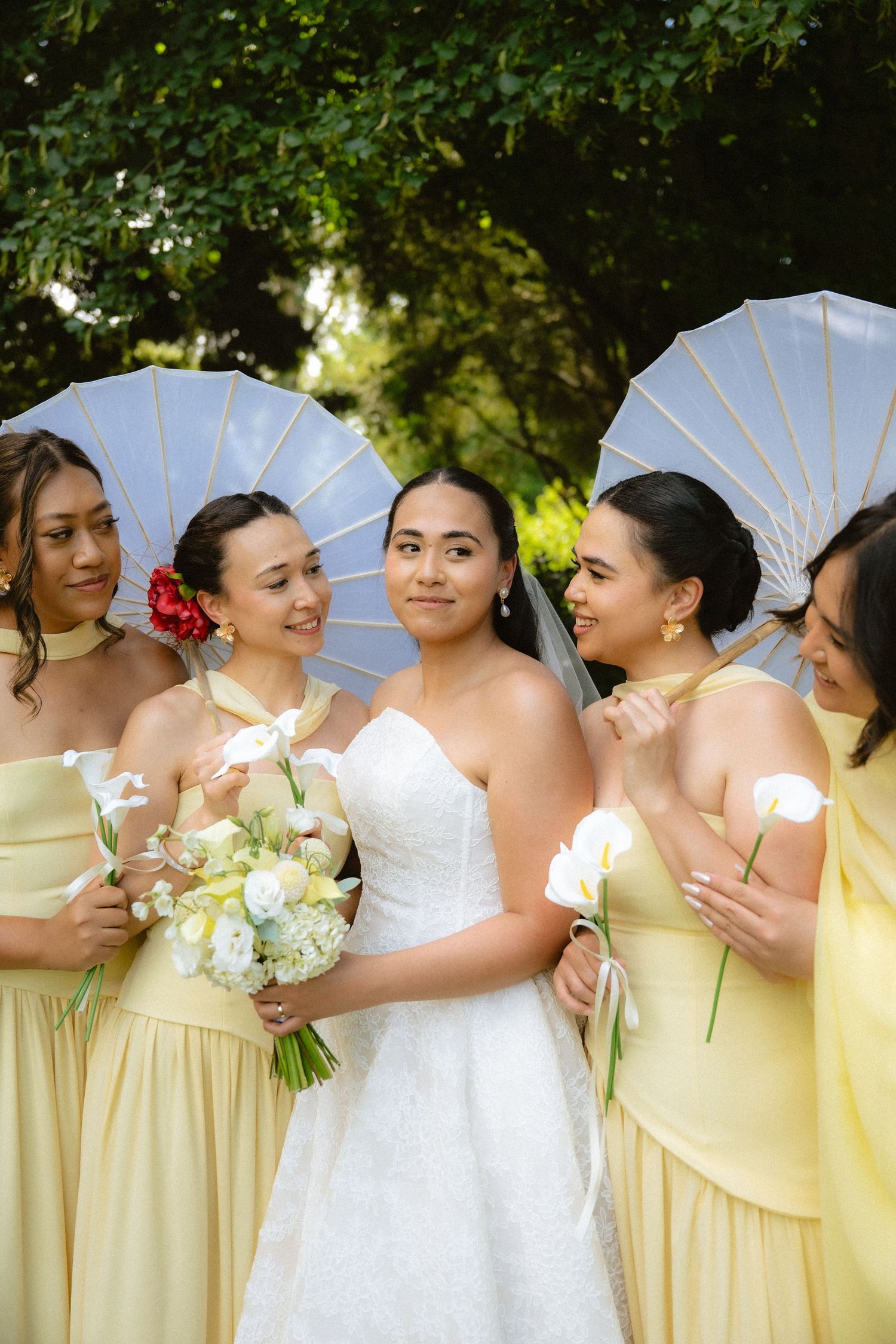 Bride in white gown holds bouquet with four bridesmaids in yellow dresses holding parasols and flowers outdoors.