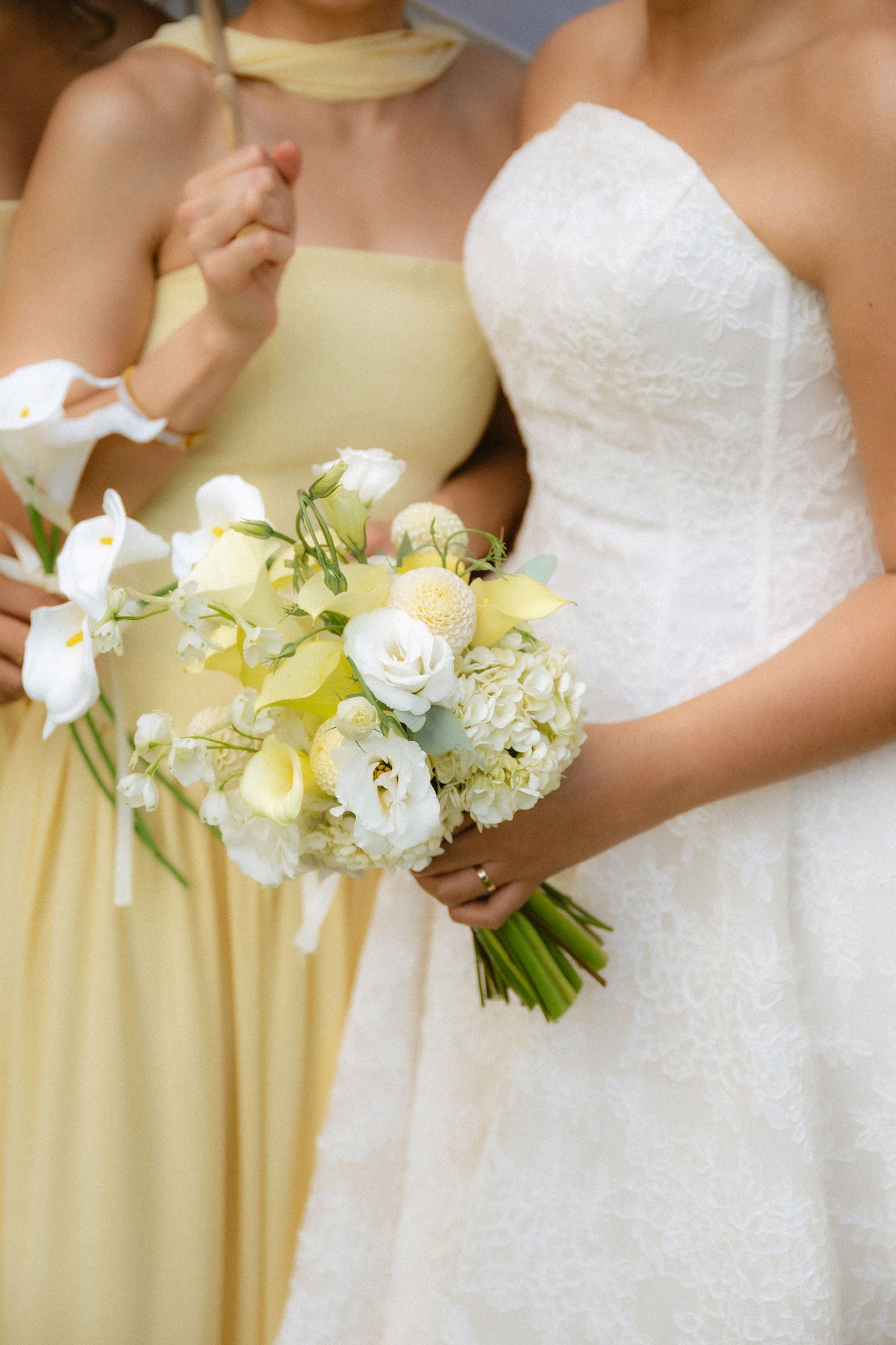 Bride in white lace gown holding yellow and white wedding bouquet beside bridesmaid in yellow dress.