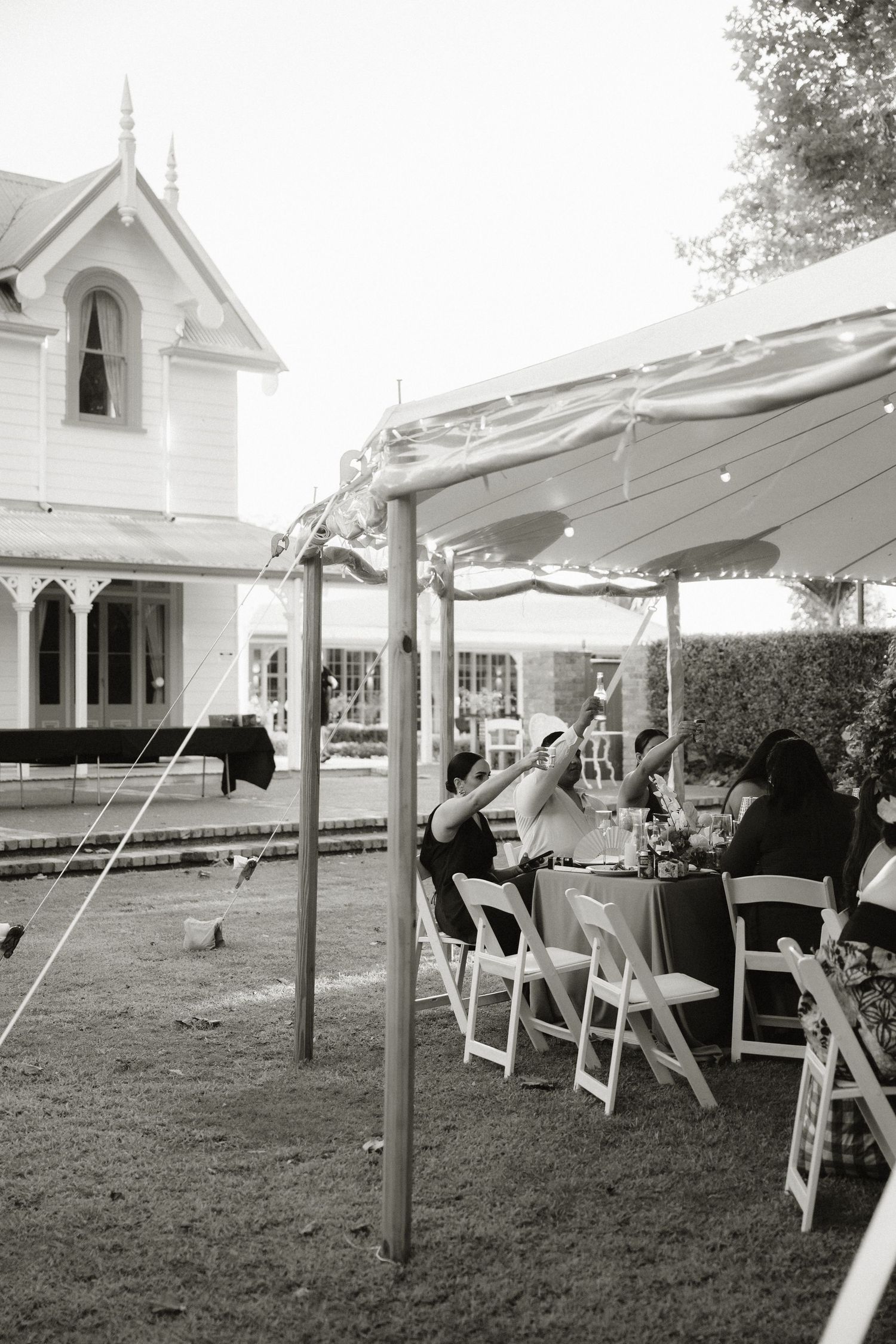 Black and white outdoor wedding reception under a tent with white folding chairs beside a Victorian house.