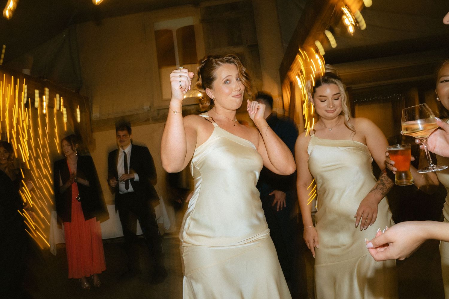 Bridesmaids in champagne satin gowns dancing joyfully at a wedding reception with sparkling lights in background.