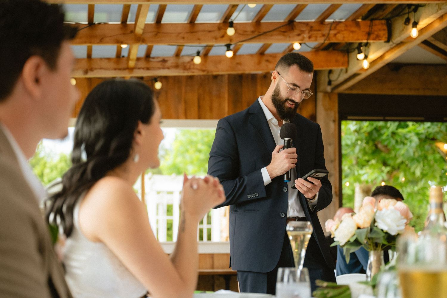 Bearded groomsman in navy suit delivers heartfelt wedding speech while holding microphone and phone at elegant outdoor reception.