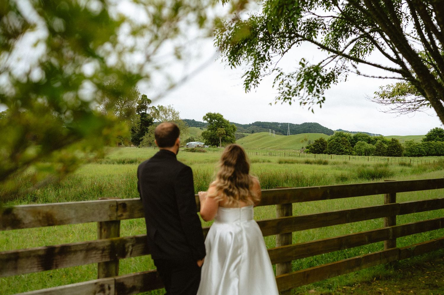 Bride and groom in wedding attire stand by a wooden fence overlooking lush green countryside fields.