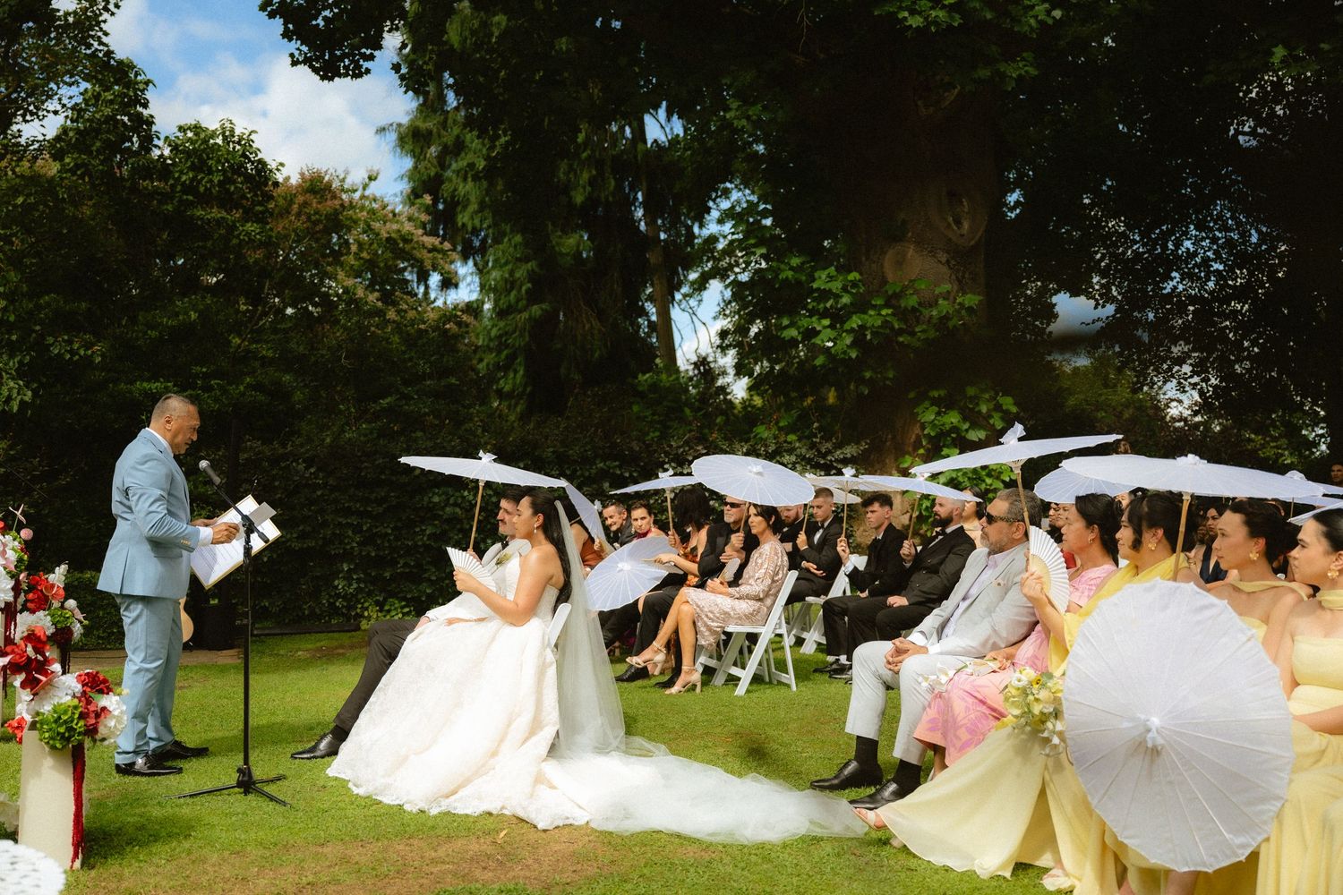 Outdoor wedding ceremony on green lawn with guests holding white parasols seated before couple and officiant under large trees.