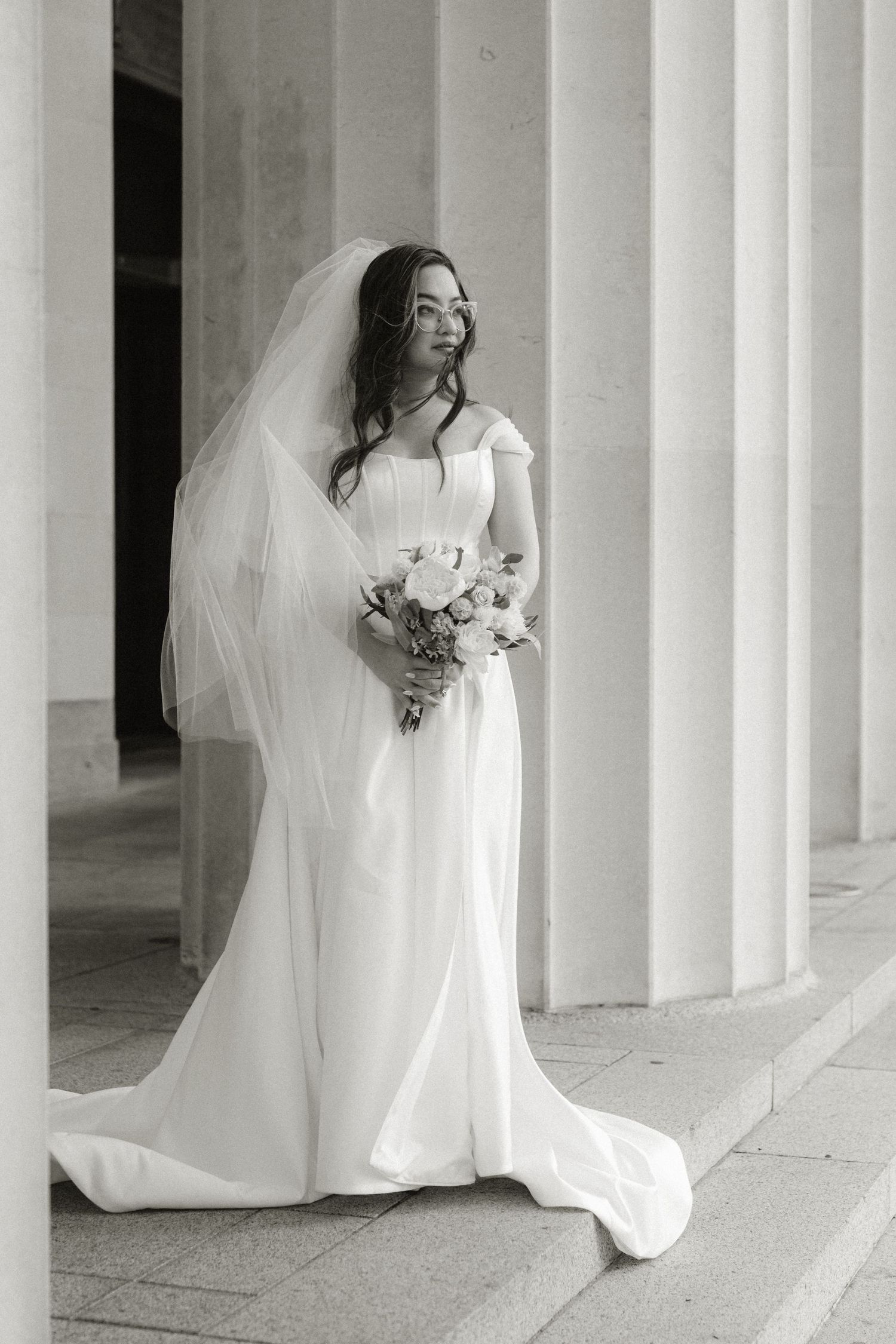 Bride in her elegant wedding gown, standing by a thick pillar, gazes thoughtfully into the distance.