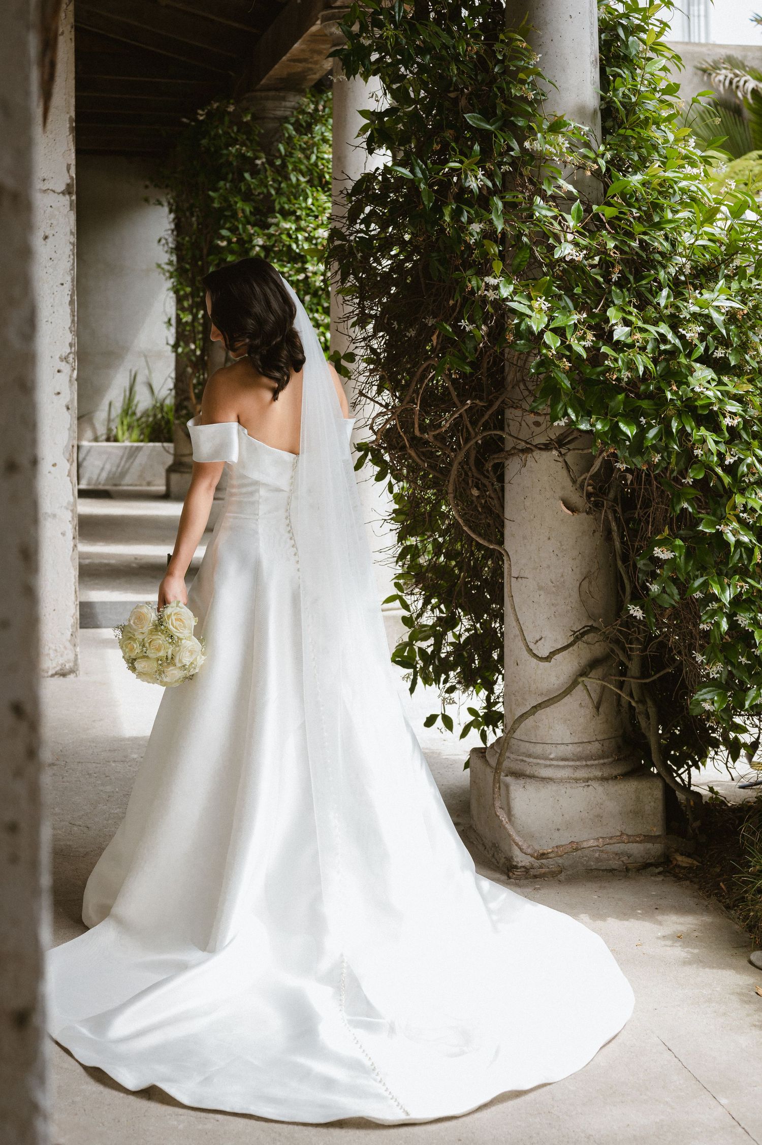 Elegant white wedding dress flows along outdoor corridor with greenery.