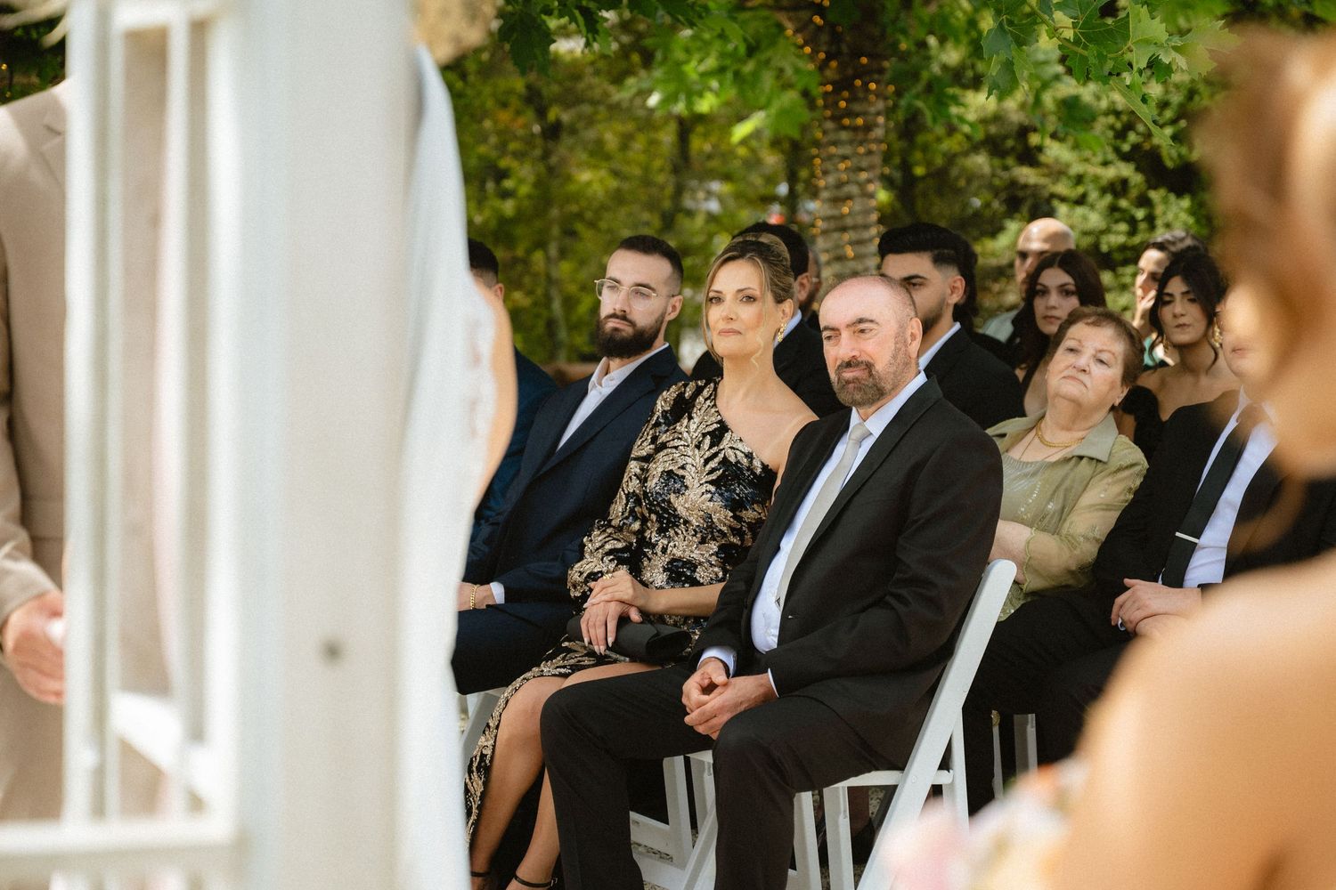 Guests seated outdoors at a wedding ceremony, smiling and watching the couple exchange vows in a garden setting.