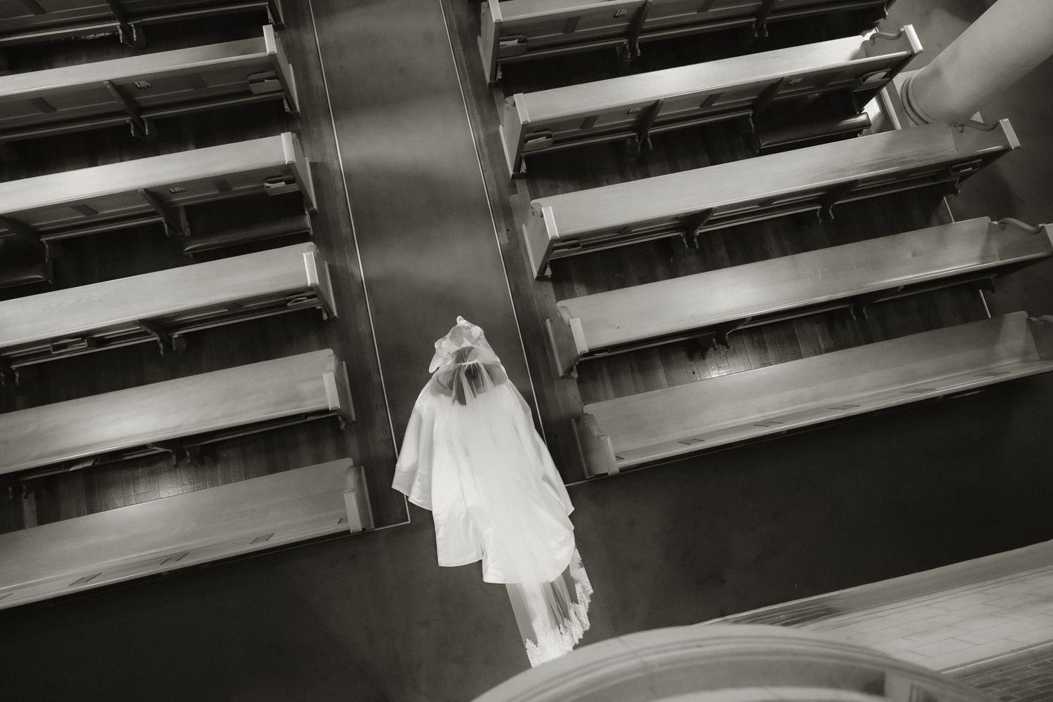 Black and white aerial view of a bride in white gown and veil walking down church aisle between wooden pews.