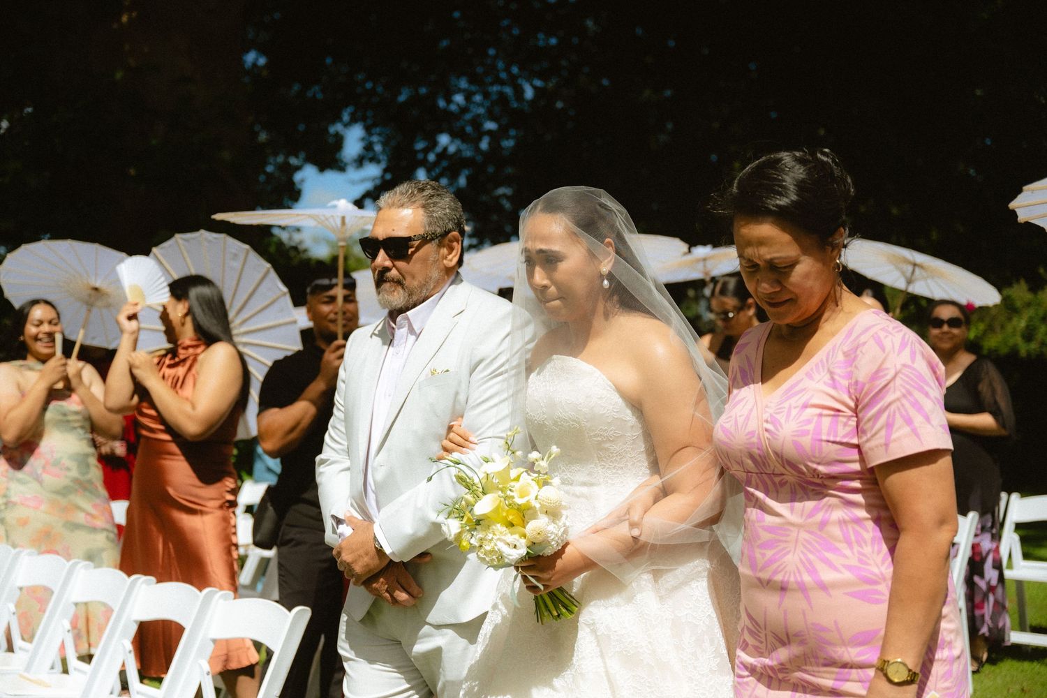 Bride walks down the aisle with family at an outdoor wedding ceremony, holding a yellow and white bouquet.