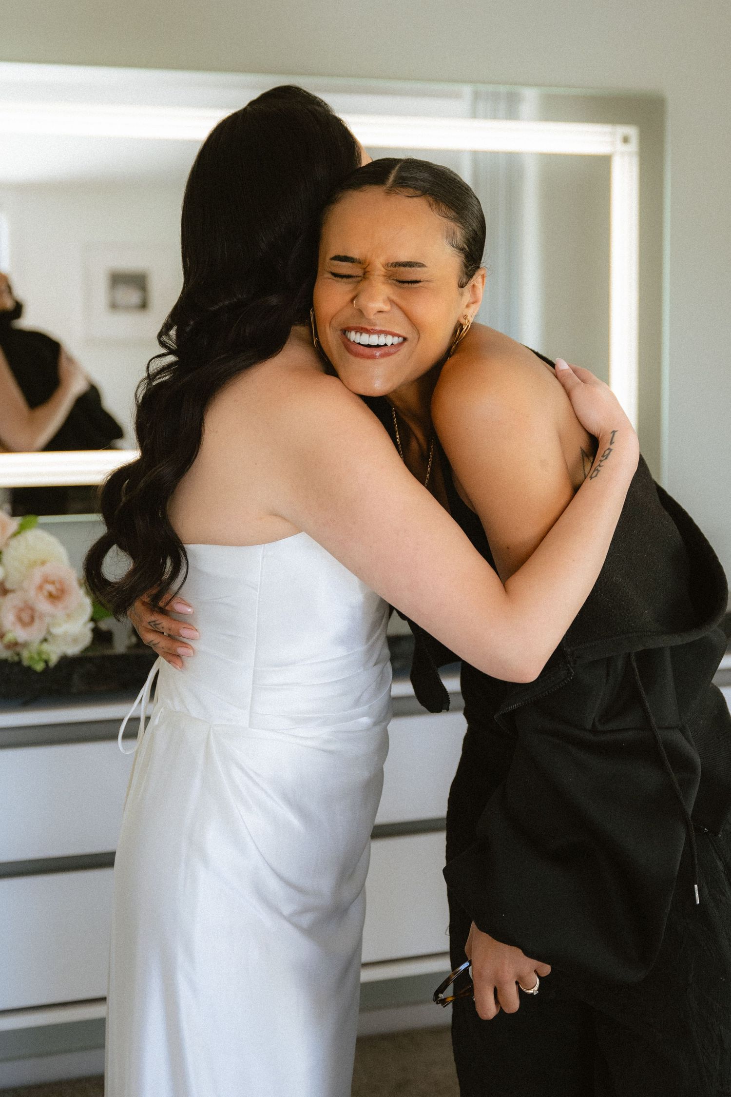 Bride in white gown shares joyful hug with smiling guest in black at wedding venue.