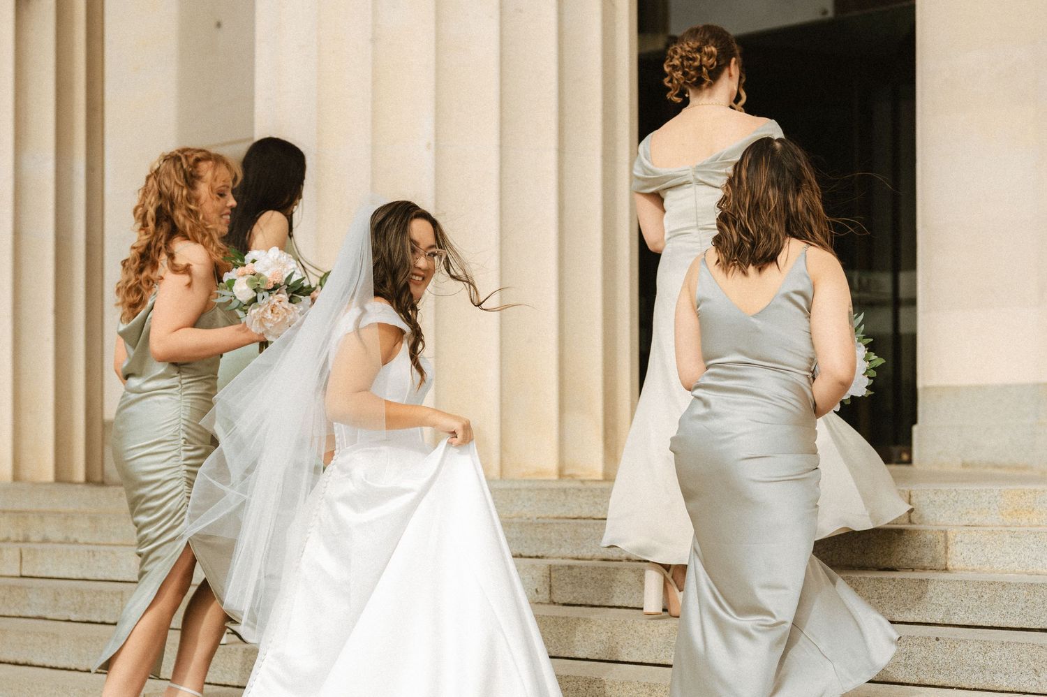 Wedding party members in gray dresses pose together near classical stone columns.