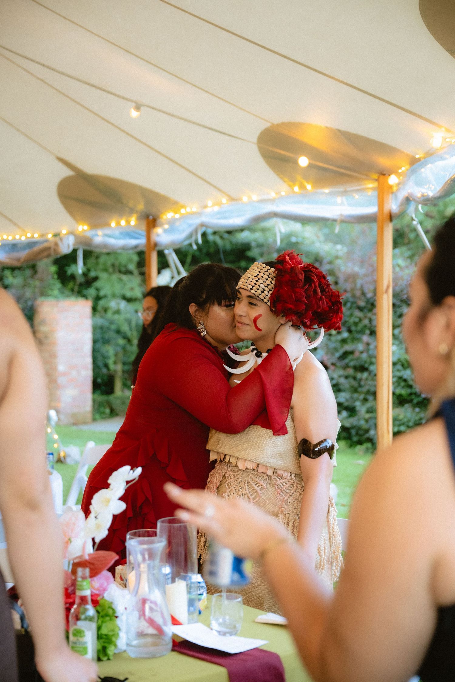 Two women in traditional attire embrace warmly under a decorated tent at an outdoor celebration.