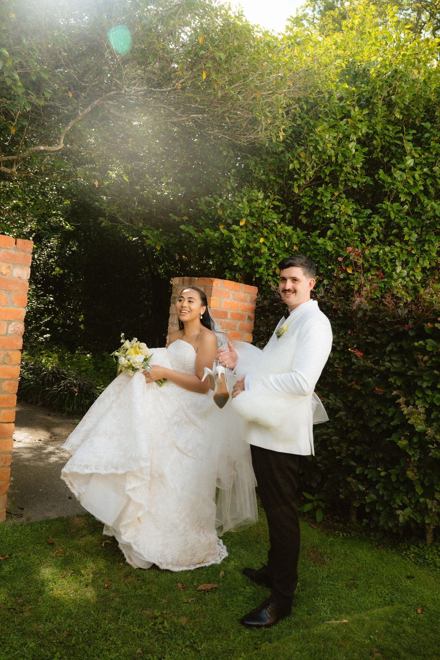 Bride in white gown and groom in white jacket hold hands, walking through a sunlit garden on their wedding day.