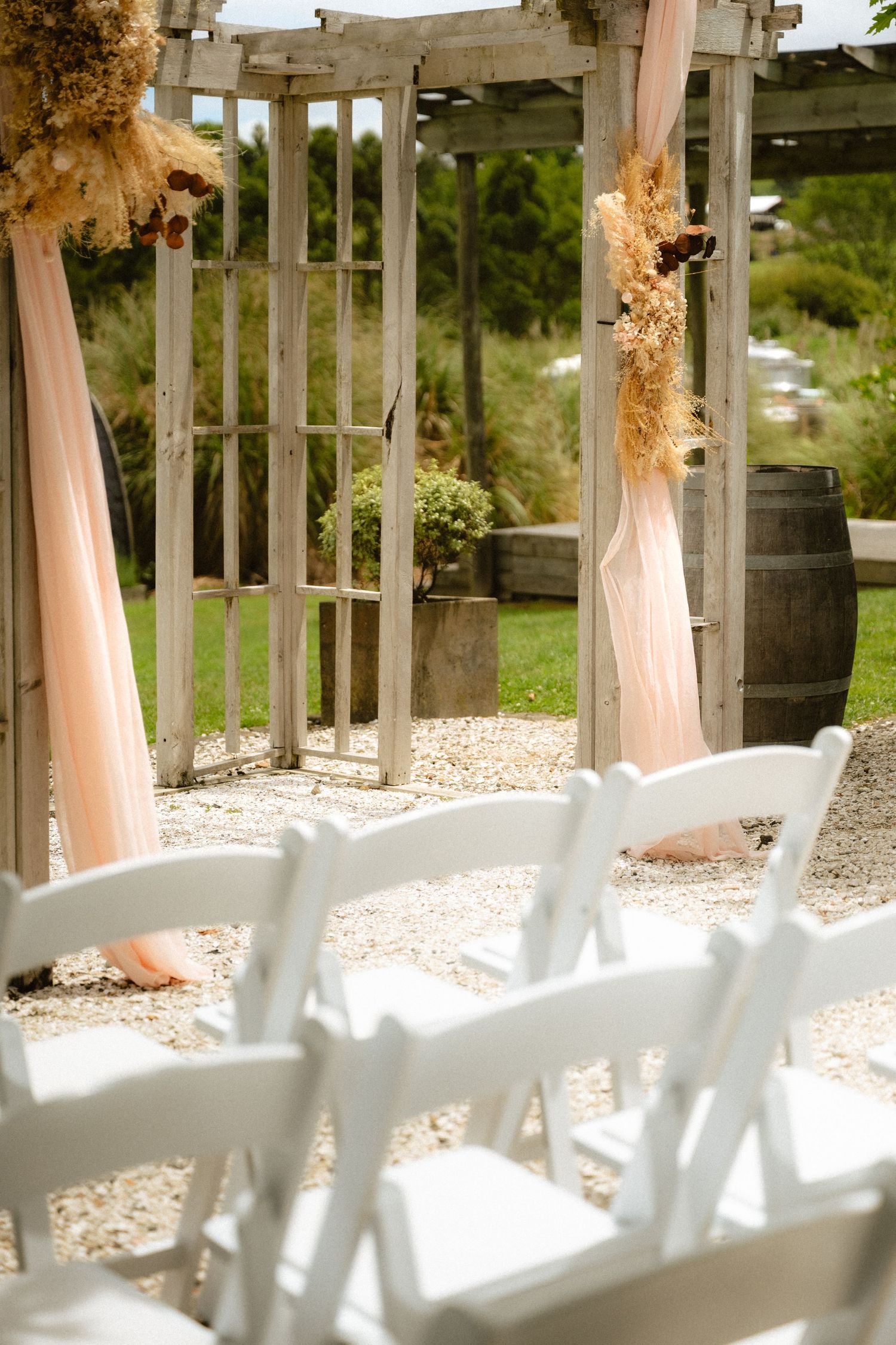 Rustic outdoor wedding ceremony setup with white chairs, wooden arbor, blush draping, dried florals, and a barrel on gravel.