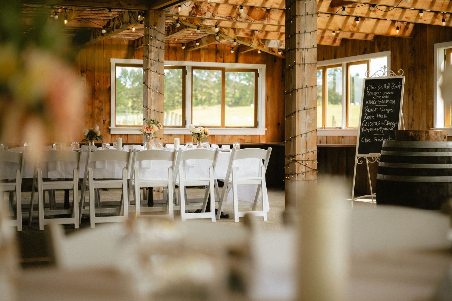 Rustic barn wedding reception with white folding chairs, string lights, and a chalkboard sign.