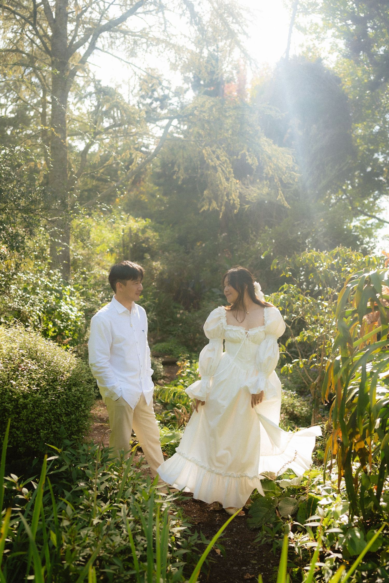 A couple walks hand in hand through a sunlit garden, the woman wearing an elegant white puff-sleeve gown.